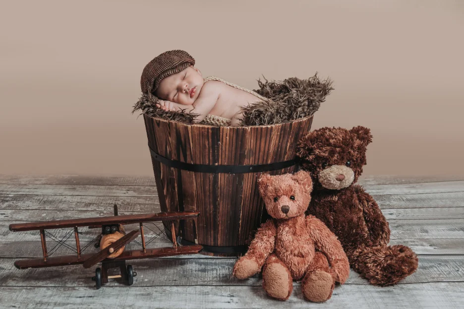 A newborn sleeping in a rustic wooden basket beside teddy bears